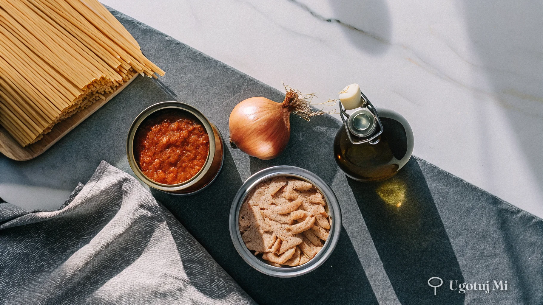 Składniki na makaron z tuńczykiem rozłożone na blacie – puszki tuńczyka, pomidory, makaron i oliwa | ugotujmi.pl